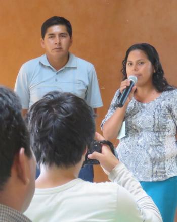 Tres personas de pie frente a un grupo, una mujer hablando por el micrófono.