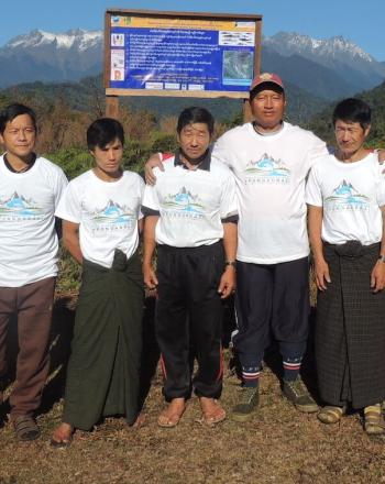 Nine people standing outside in front of sign, mountains in background.