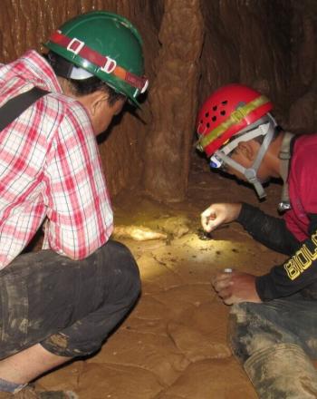 Two men wearing hard hats inside a cave, looking at specimen.
