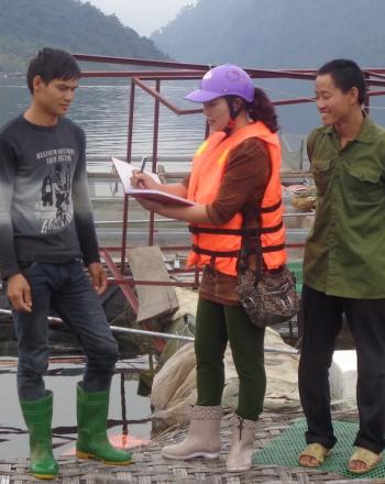 Junto al agua, dos hombres se paran junto a una mujer que está escribiendo en un cuaderno.