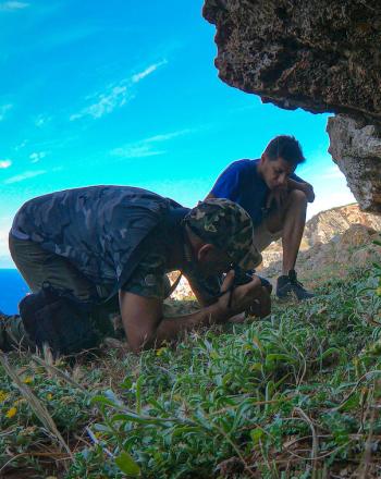 Dos hombres en una colina, muy por encima del agua, uno está inclinado observando al otro fotografiando una planta de cerca.