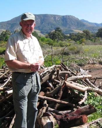 Man standing outside, smiling at camera. Pile of branches behind him, mountain in the background.