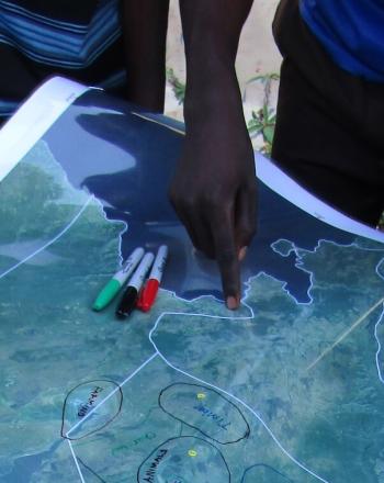 Map on table, a few people's hands holding down map, one pointing toward location..
