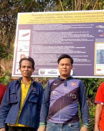 Four men standing in front of large signboard, forest behind it.