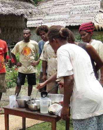 A group of men and women encircle a woman at a small table demonstrate coconut oil pressing.