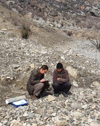 Two men in rocky landscape crouched on ground, talking.