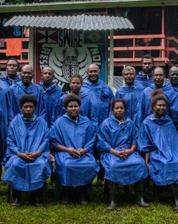 Grupo de unas 20 personas con ponchos azules para la lluvia, de pie/sentados frente a la estación de guardabosques.