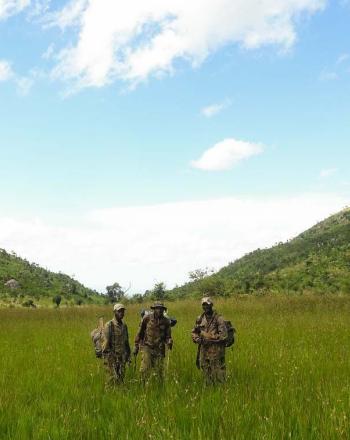 Three men standing in field, hills is background.