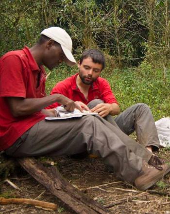 Two men sitting on forest floor, looking at notebook.
