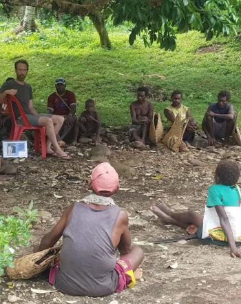 About 20 people sitting outside in a circle, under a tree.