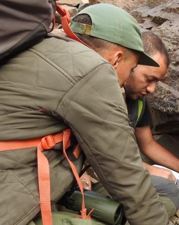 Two men crouching down near boulders, examining papers.
