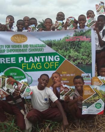 Group of boys stand around tree planting sign, outside.