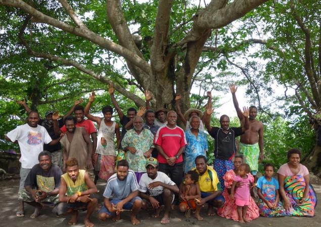 Several men, women and children gather and wave for a group photo in front of a large tree in Tafea Province, Vanuatu.