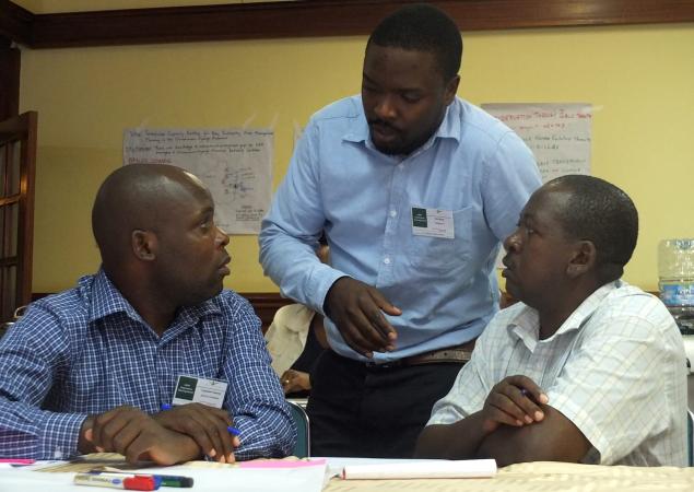 Three men talking, two seated and one standing.