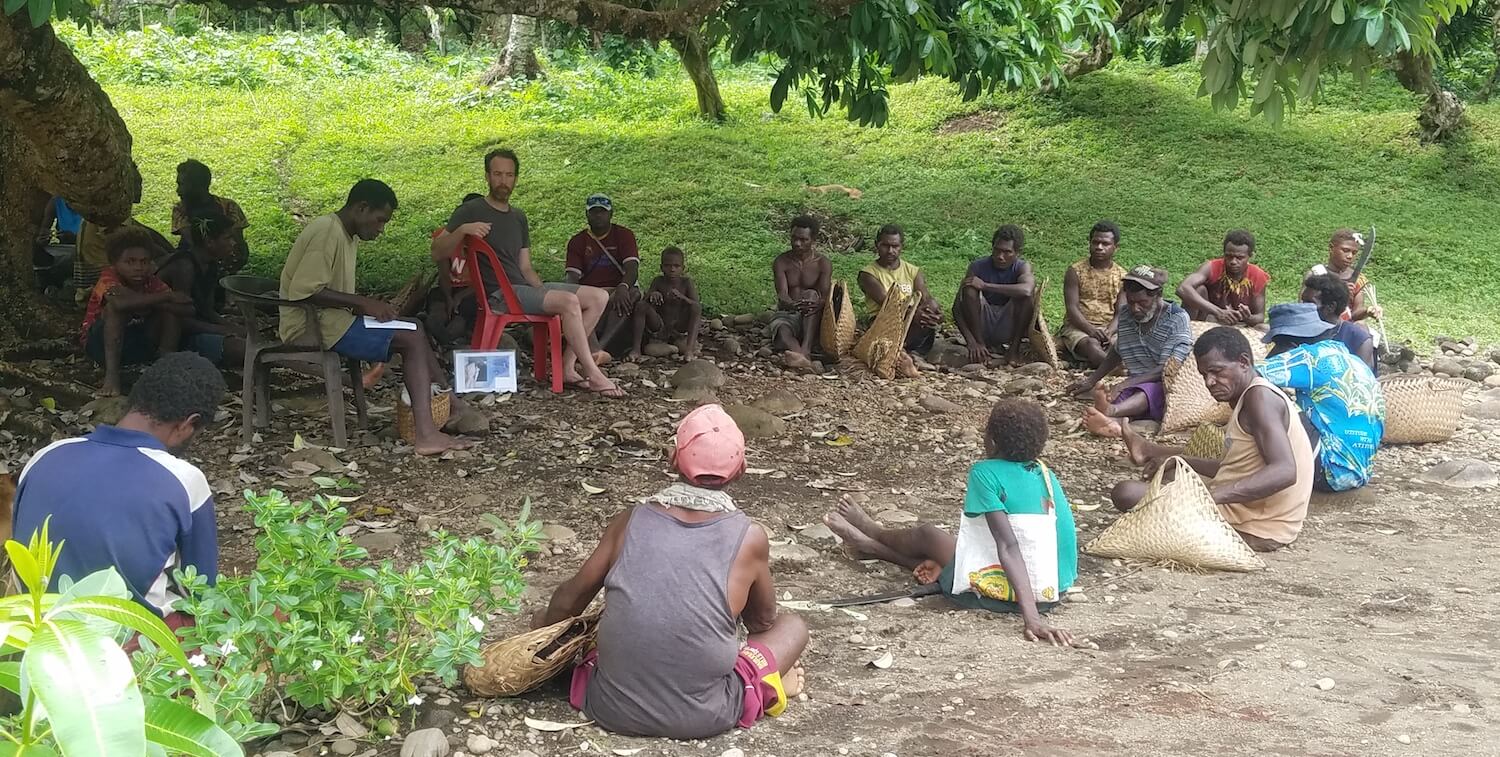 About 20 people sitting outside in a circle, under a tree.