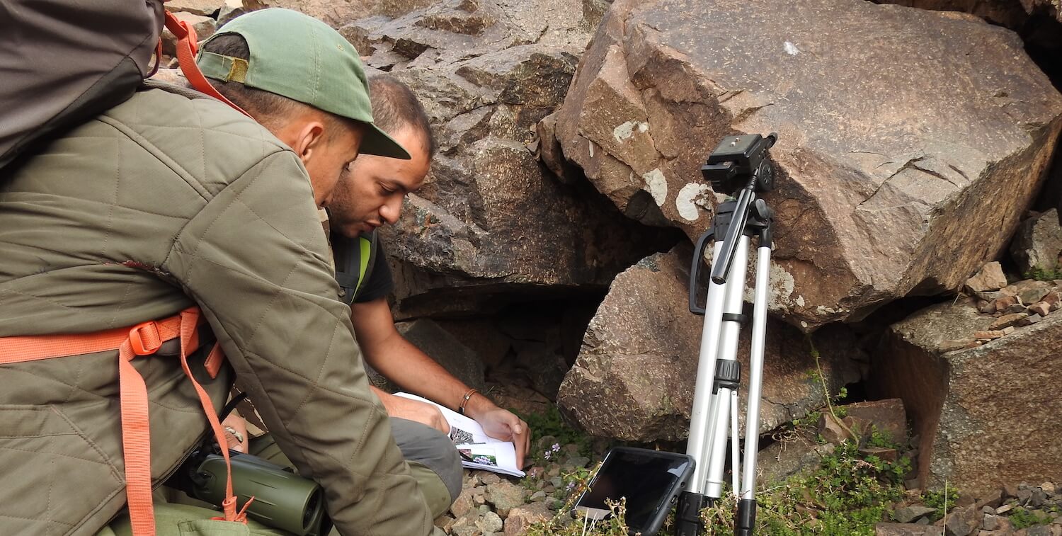 Two men crouching down near boulders, examining papers.