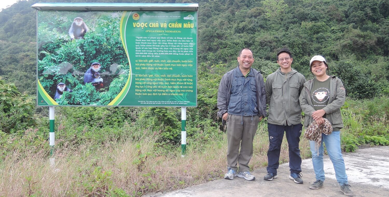 Trang standing with 2 men next to large sign with writing and photos of monkeys.