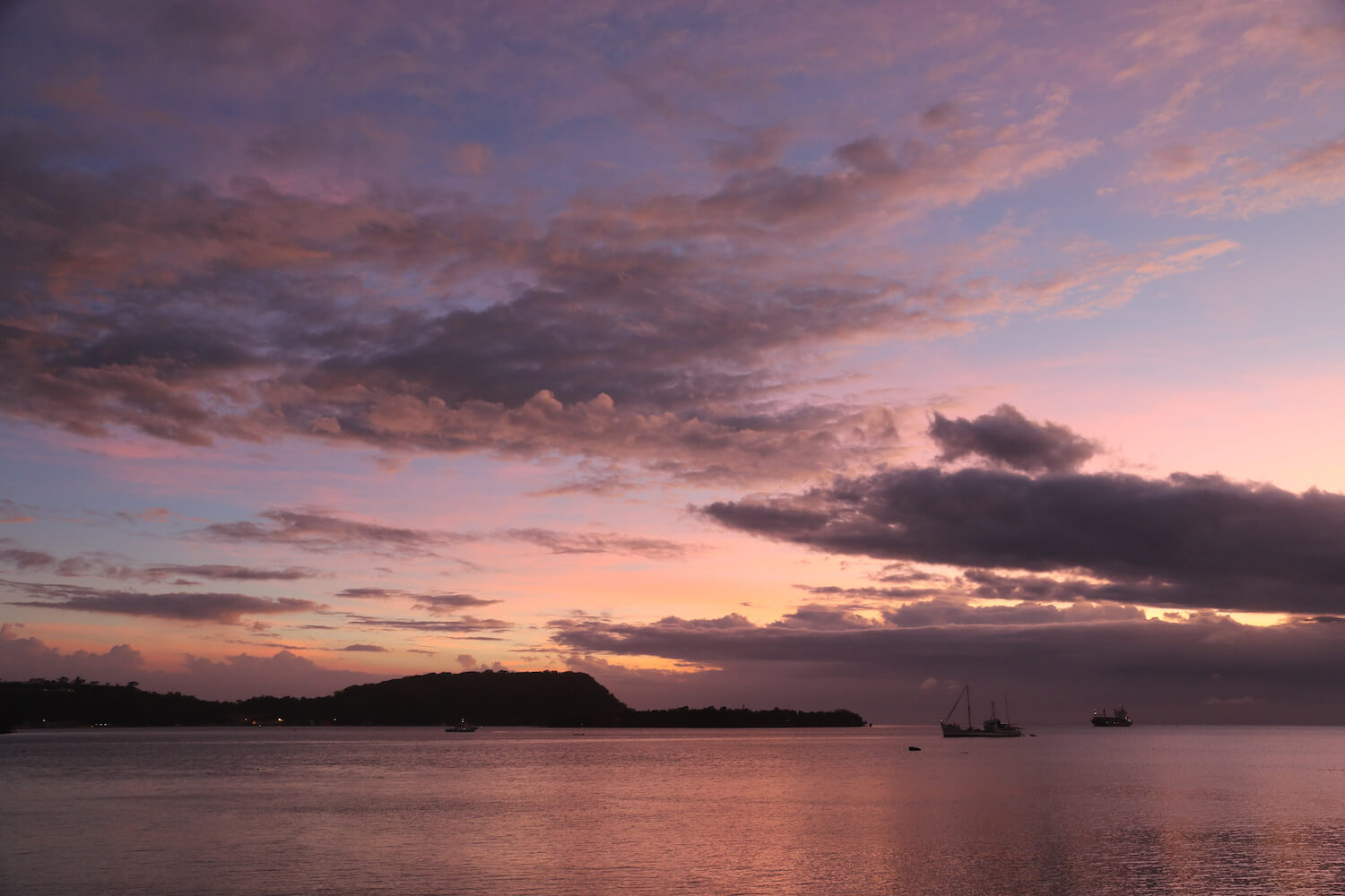 Puesta de sol sobre el océano, algunos barcos e isla con montaña en la distancia.