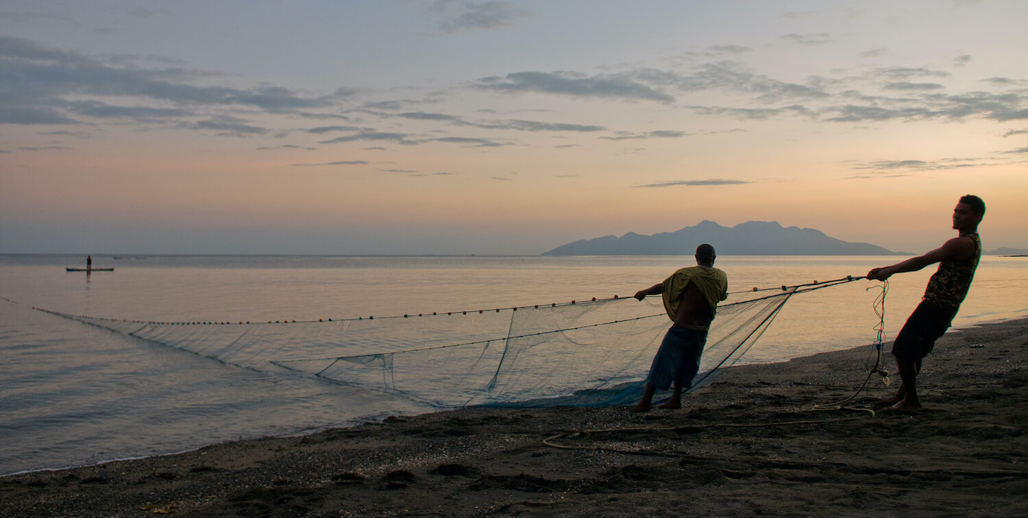 Two fishermen pulling in a net from the ocean at sunset.