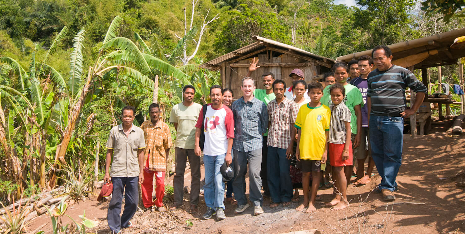 A large group of men, women and children stand outside of a small wooden building with a forested area in the background.