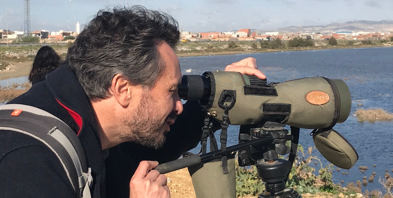 Close-up of man looking through a monocular, water behind him and buildings in background.