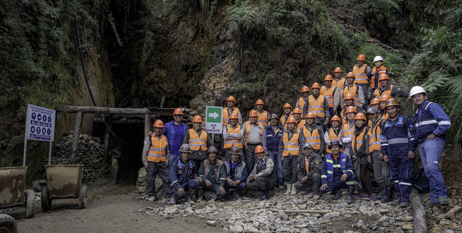 ほとんどがオレンジ色のベストとヘルメットをかぶった約25人の鉱山労働者のグループが、鉱山の外に立っています。