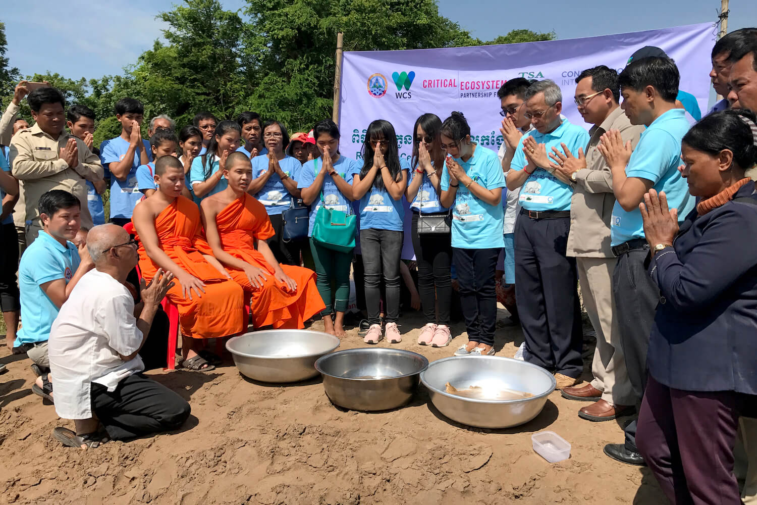 Group of people, including two monks in orange robes, surround three bowls, and pray.