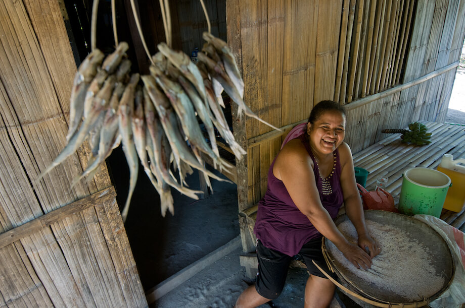 A seated woman smiles while looking up as she sifts rice in a woven container.