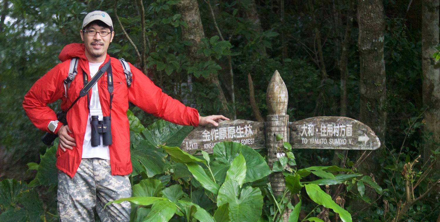 Un hombre se para al lado de una señal de sendero en un bosque.