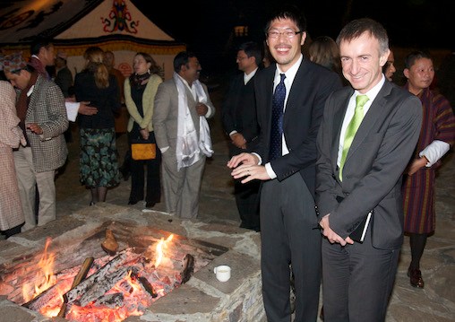 Two men stand near fire pit with several people in the background.